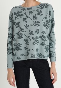 Ljusblå tröja med ett texturerat bladmönster i mörkgrönt. Rund halsringning och långärmade, kombinerad med mörka jeans.