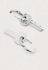 Dvoje stříbrných náušnic ve tvaru klíče na bílém pozadí, každé s zakřiveným zapínáním.