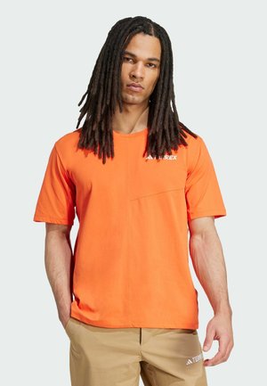 Man met lange dreadlocks, gekleed in een oranje T-shirt en beige broek met het merk "TERREX", staand tegen een effen lichte achtergrond.