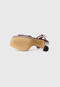Burgundy syntetisk högklackad sandal med ankelrem, en slät glänsande yta och en räfflad beige gummisula med svarta detaljer.