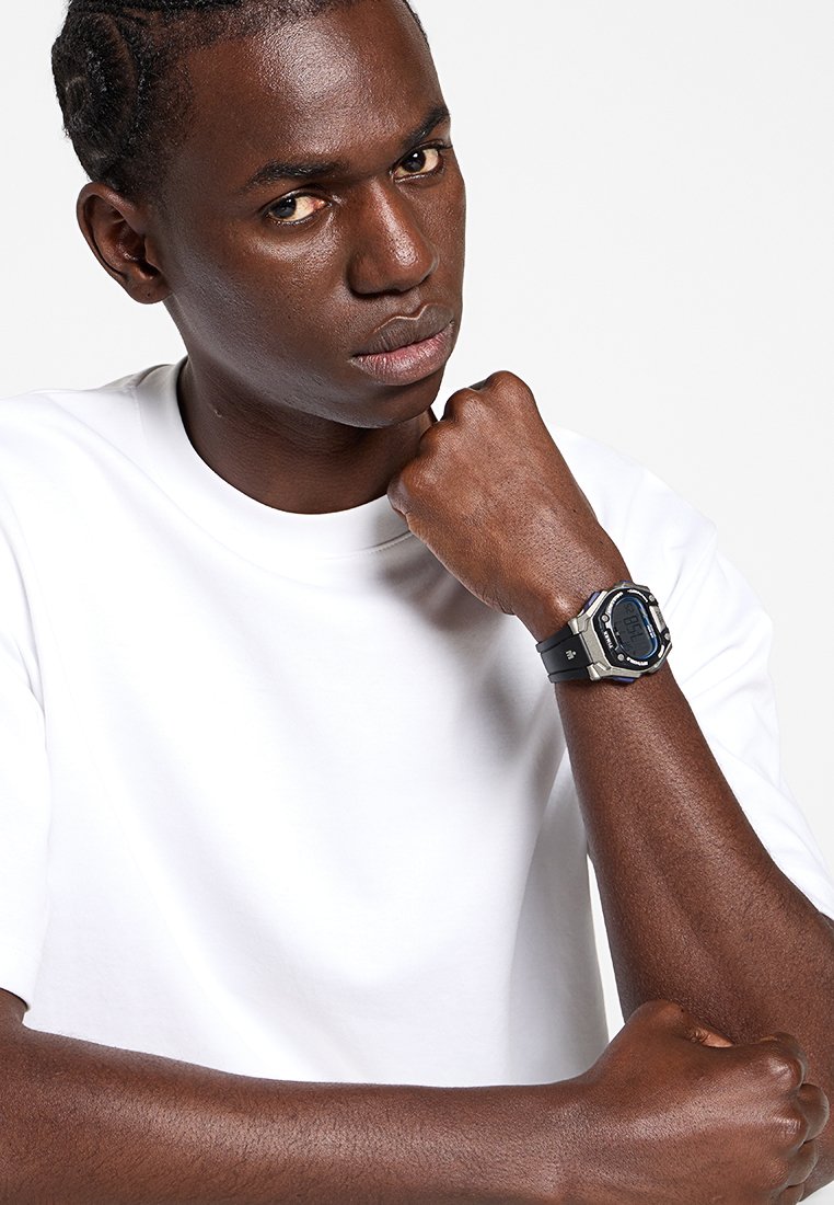 Reloj digital con una correa de goma negra, acentos en tono plateado, cara rectangular y una pantalla digital azul. Llevarlo en una muñeca con piel morena.
