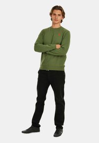 Green sweatshirt with ribbed cuffs and hem, featuring a small red logo. Paired with black jeans and black sneakers.