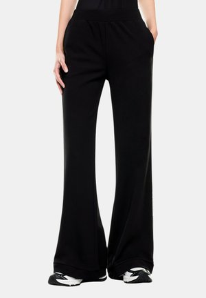 Pantaloni neri a gamba larga indossati con scarpe da ginnastica nere e bianche, mani appoggiate casualmente nelle tasche, mostrati dalla vita ai piedi.