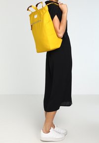 Zaino giallo con forma rettangolare, realizzato in materiale resistente. Presenta due spallacci in pelle marrone e una tasca frontale con zip.