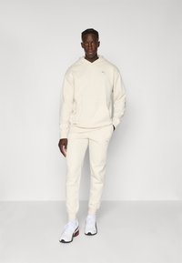 Cremefarvet hættetrøje med en frontlomme og en diskret logo, parret med matchende tapered joggers. Sættet har et struktureret stof.