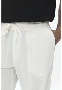 Białe lekkie spodnie z wiązaniem w pasie i bocznymi kieszeniami. Materiał ma teksturowaną powierzchnię z subtelnymi liniami i luźnym krojem.