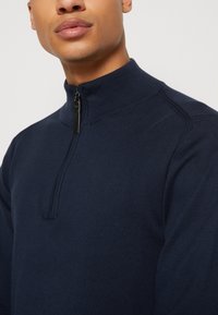 Suéter de cuello alto con cremallera en color azul marino, elaborado en una suave tela de punto, con un cuello levantado y costuras sutiles. La cremallera tiene un tirador negro.
