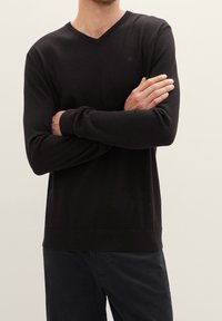 Maglione nero lavorato a maglia con scollo a V, maniche lunghe e polsini a costine. Presenta un logo sul petto. Tessuto liscio con design aderente.