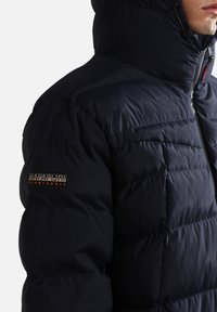 Marineblå pufferjakke med høj krave, quiltet struktur og et fremtrædende Napapijri-logo på ærmet. Har lynlås lukning.