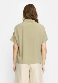 Light olive green short-sleeved blouse with a collar, soft texture, and a relaxed fit, ending at the waist, paired with beige trousers.