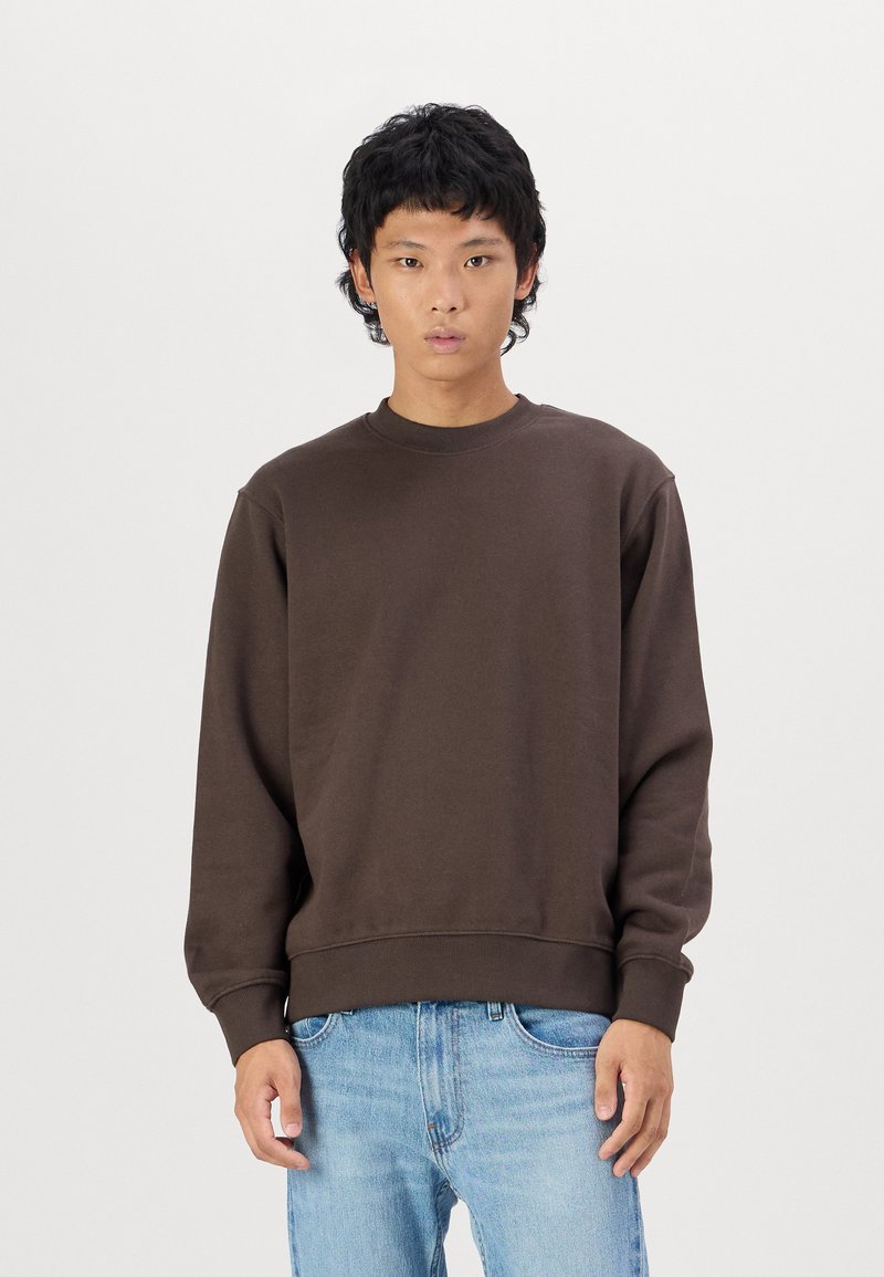 Felpa marrone con colletto tondo, polsini e orlo a costine. Indossata con jeans azzurri chiari, presenta una texture liscia e una vestibilità comoda.