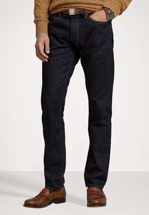 Vaqueros de mezclilla azul oscuro con costuras en contraste, corte Slim, diseño de cinco bolsillos y un cinturón de cuero marrón. Combinados con zapatos mocasines marrones.