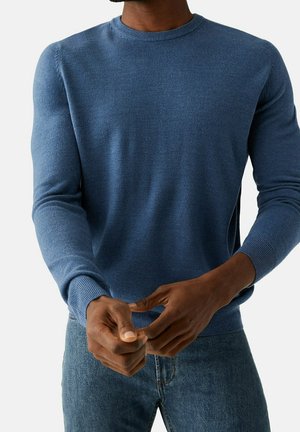 Uomo che indossa un maglione blu girocollo e jeans blu, mentre sistema la manica con entrambe le mani su sfondo bianco.