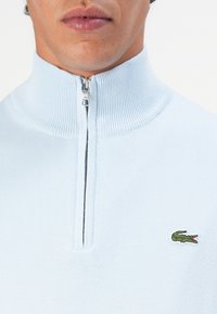 Svetlo modra zip-up jopica iz mehkih materialov. Ima rebrast ovratnik in majhno zeleno Lacoste logo na prsih.