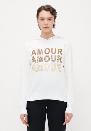 Person som har på seg en hvit hettegenser med "AMOUR" trykket tre ganger i beige, leopardmønster, og lys beige, kombinert med svarte bukser.