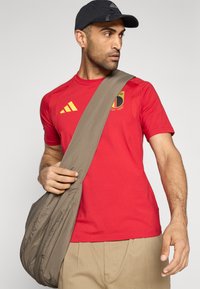 Rdeča športna majica z rumenimi poudarki in belgijskim grbom. Oseba nosi temno kapo in drži bež torbo čez ramo. Vidne so kaki hlače.