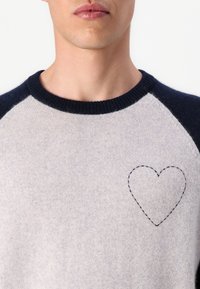 Homme portant un pull gris clair avec un col bleu marine foncé et un contour de cœur brodé sur le côté gauche de la poitrine.