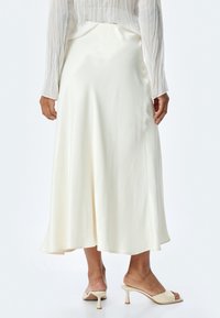 Jupe midi en satin ivoire avec une forme fluide. Texture lisse, fente sur le côté et une légère brillance. Associée à des mules à talons ouverts beiges.