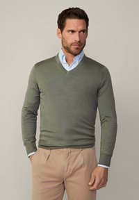 Pull en tricot à col en V de couleur vert olive, manches longues, poignets côtelés, associé à une chemise bleu clair et un pantalon beige.