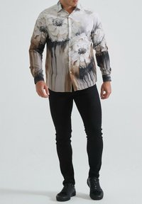 Chemise à boutons avec un imprimé floral dans des tons crème et marron, présentant des accents noirs aux poignets. Associée à un pantalon slim noir et des baskets sombres.