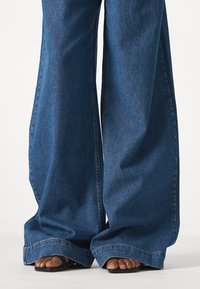 Široké džíny z denimu v tmavě modré barvě, s vysokým pasem, roztrhaným spodním lemem a hladkou texturou. Černé ploché sandály jsou částečně viditelné.