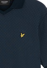 Marinblå texturerad polotröja med V-ringning; har en liten gul broderad örnlogotyp på vänster bröst.