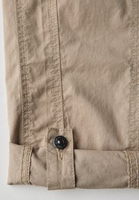 Beige bavlněná tkanina s mírně zmačkanou strukturou, se kontrastním šitím a černým knoflíkem na manžetě.