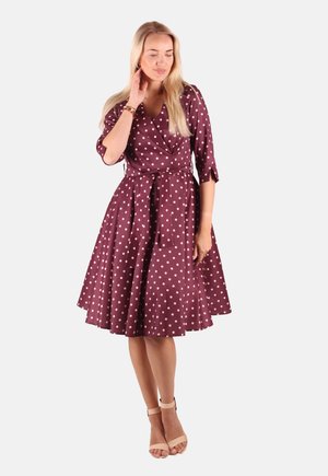 Frau mit langen blonden Haaren, die ein knielanges dunkelrotes Kleid mit weißen Punkten und beigefarbenen Pumps mit Knöchelriemen trägt, steht vor einem schlichten Hintergrund.