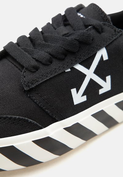 Μαύρο αθλητικό παπούτσι με παχιά μαύρα κορδόνια, λευκό λογότυπο με σταυρωτά βέλη στο πλάι και λευκή σόλα με μαύρες διαγώνιες ρίγες.