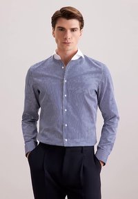 Gestreept shirt in blauw-witte katoen met een subtiele textuur, voorzien van een opstaande kraag en knopen aan de voorkant. Draagt bijpassende donkere broek.