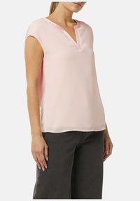Jasnoróżowa bluzka z krótkim rękawem i dekoltem w kształcie litery V, wykonana z gładkiego materiału, charakteryzująca się luźnym krojem i lekko zaokrąglonym dołem.