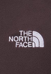 Tessuto in fleece marrone con un logo ricamato bianco di "THE NORTH FACE" caratterizzato da un design semicircolare, che enfatizza la texture e il branding.