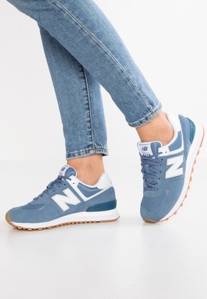 Person wearing blue and white New Balance sneakers paired with light blue denim jeans, standing on a white floor.