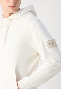 Hoodie Calvin Klein v krémovej farbe, s klokankovým vreckom, rebrovanými manžetami a vyšitým logom na rukáve. Jemná, hladká textúra.