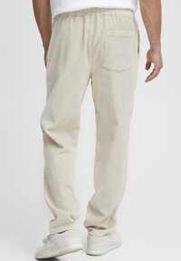 Pantalon en velours côtelé beige avec une taille élastique, une poche arrière et une coupe ample, associé à des baskets blanches. Texture côtelée verticale visible.