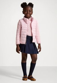 En ung jente står iført en rosa quiltet jakke over en rosa genser, en marineblå plisserte skjørt, marineblå knelengde sokker, og brune sko.