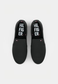 Paire de chaussures noires sans lacets avec un petit logo Tommy Hilfiger sur le dessus et un marquage blanc visible à l’intérieur des semelles.