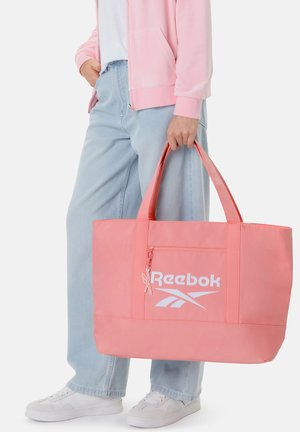 Person trägt hellblaue Jeans, weiße Turnschuhe, eine rosa Jacke und hält eine große rosa Reebok-Tasche an den Griffen.