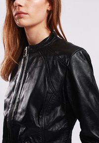 Giubbotto di pelle nera con colletto alto, chiusura con zip e dettagli di cucitura sottili. Texture liscia con un design aderente.