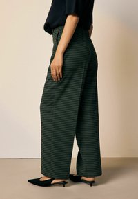 Grüne kariierte weite Hose mit elastischem Bund. Das Modell trägt schwarze Absatzmules. Einfacher Hintergrund, Seitenansicht, die die Textur des Stoffes zeigt.