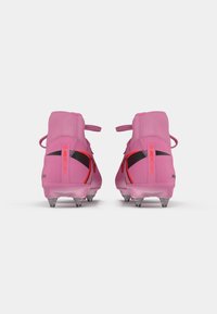 Roze voetbalschoenen met een slank ontwerp, voorzien van een hoge enkel, zwarte accenten en doorzichtige noppen voor grip op grasoppervlakken.