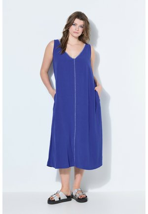 Kvinde står med hænderne i lommerne iført en ærmeløs blå midikjole og sølvfarvede åbne sandaler mod en ensfarvet baggrund.