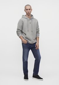 Jeune homme debout les mains dans les poches, portant un sweat à capuche gris clair, un jean bleu et des baskets noires sur un fond blanc uni.