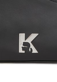 Musta nahkatägi, jossa on hopeinen K-logo profiilimuotokuvalla. Pinta on sileä ja siinä on hienovarainen tekstuuri.