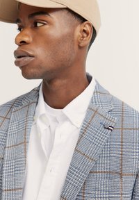 Hellblau kariertes Blazer mit braunen Mustern, strukturiertem Stoff, geknöpftem Revers und einer Anstecknadel. Getragen über einem weißen Hemd mit Kragen und einer beigen Mütze.