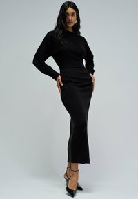 Vestido de punto negro con un cuerpo ajustado, mangas largas y un dobladillo acampanado. Combinado con estilettos negros de charol y un diseño minimalista.