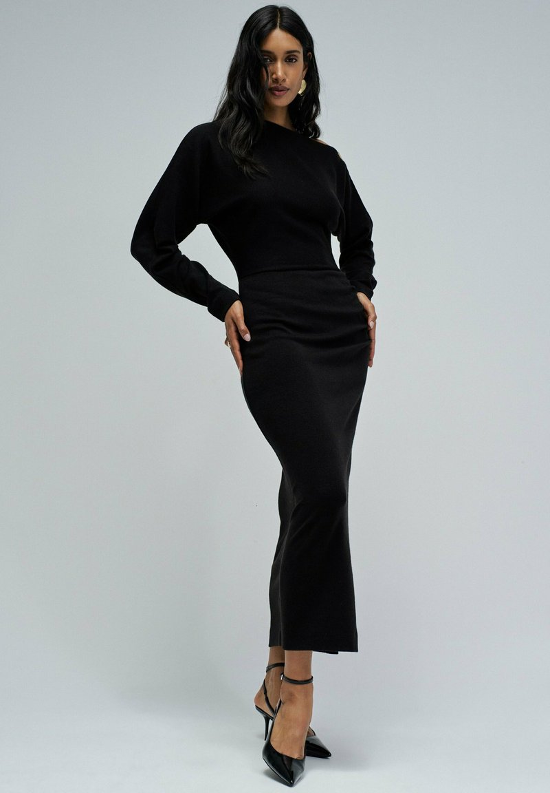 Vestido de punto negro con un cuerpo ajustado, mangas largas y un dobladillo acampanado. Combinado con estilettos negros de charol y un diseño minimalista.