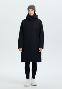 Cappotto lungo nero con cappuccio, chiusura con zip e tasche laterali. Indossato sopra leggings neri e sneaker bianche, abbinato a un berretto marrone.