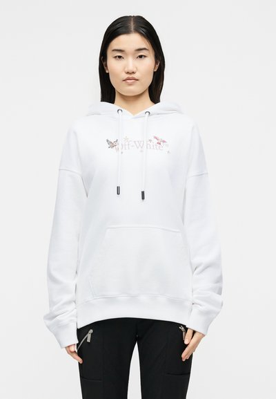 Γυναίκα που φοράει λευκό φούτερ Off-White με σχέδια πουλιών και μαύρο παντελόνι με τσέπες με φερμουάρ, στέκεται μπροστά σε απλό φωτεινό φόντο.