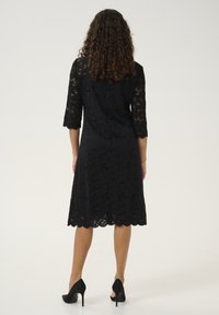 Black lace dress with three-quarter sleeves, knee-length hem, scalloped edges, and floral pattern. Worn with black high-heeled shoes.